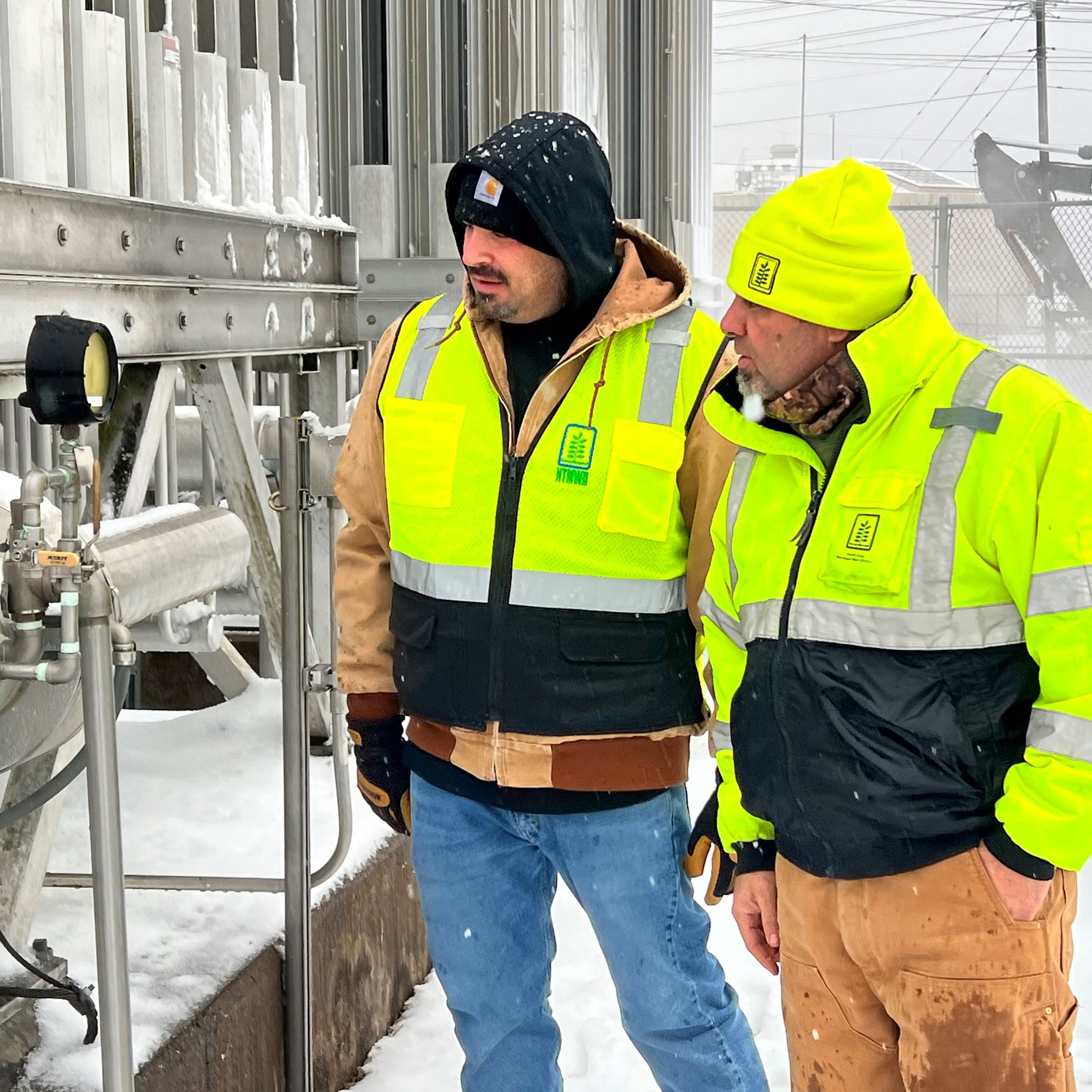Two men in safety jackets look at water gauges outside in the snow.