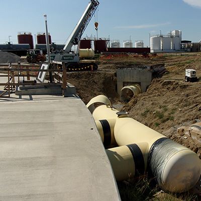 An 8-foot pipe is on the ground before being connected to another pipe. There is a crane and tanks.